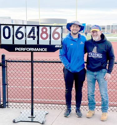 UNK JAVELINTHROWER RYLAN BASART, left, along with his dad, Justin stand beside the record-setting distance sign at the Central Nebraska Challege last Saturday. His toss of 64.86 meters (212’9”) broke the record which had stood for 32 years. (Courtesy Photo)